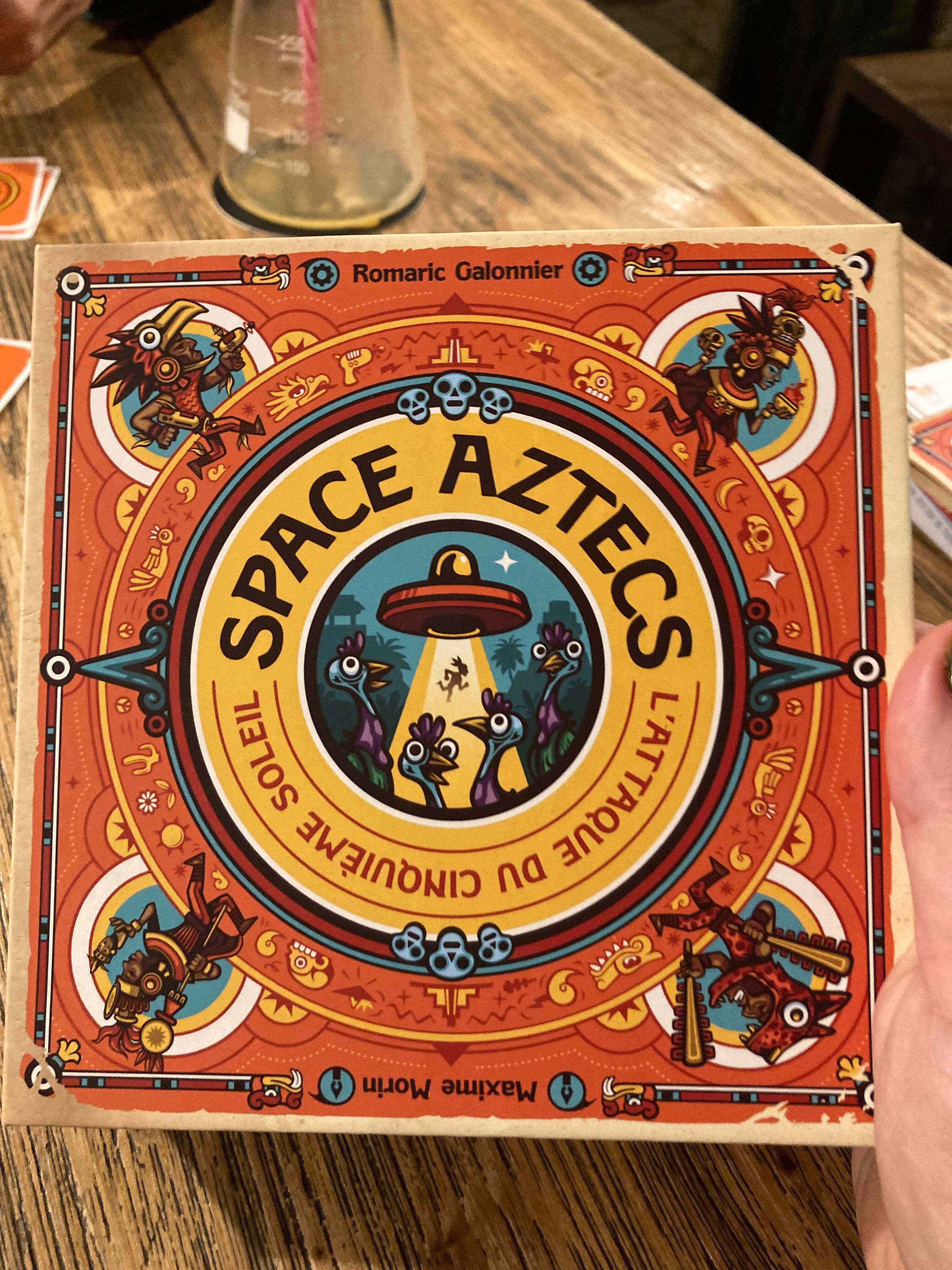 Boîte du jeu Space Aztecs, avec des illustrations de couleur vive représentant des guerriers aztèques, des dindes et une soucoupe volante en train d'enlever quelqu'un. L'illustration joue sur la symétrie et des motifs type aztèque stylisés. Le sous-titre du jeu est "L'attaque du cinquième soleil", et les noms des deux créateurs sont Romaric Galonnier et Maxime Morin.