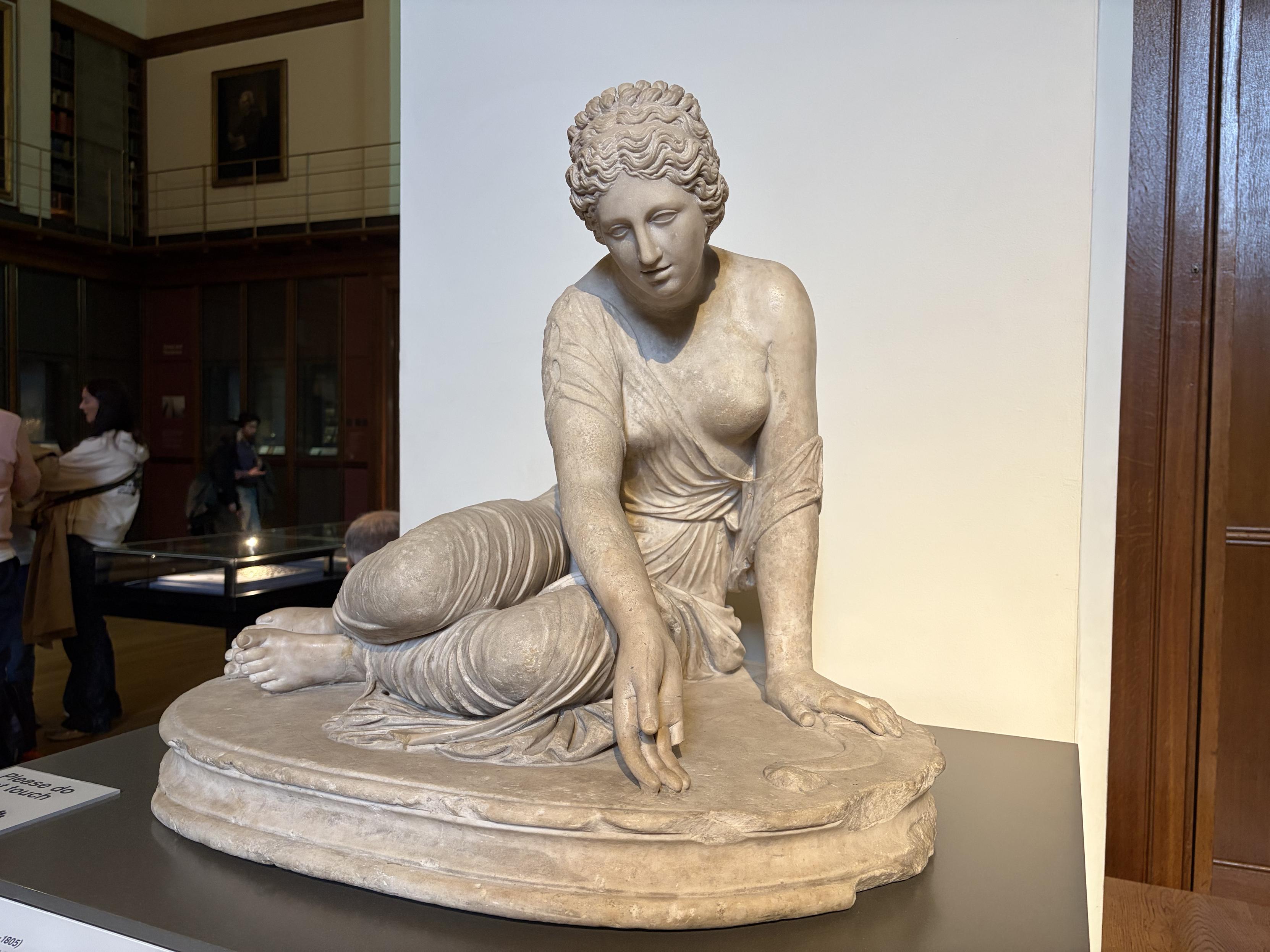 Sculpture d&rsquo;une femme assise par terre et qui d&eacute;signe le sol avec sa main. Elle a une expression passablement saoul&eacute;e.
