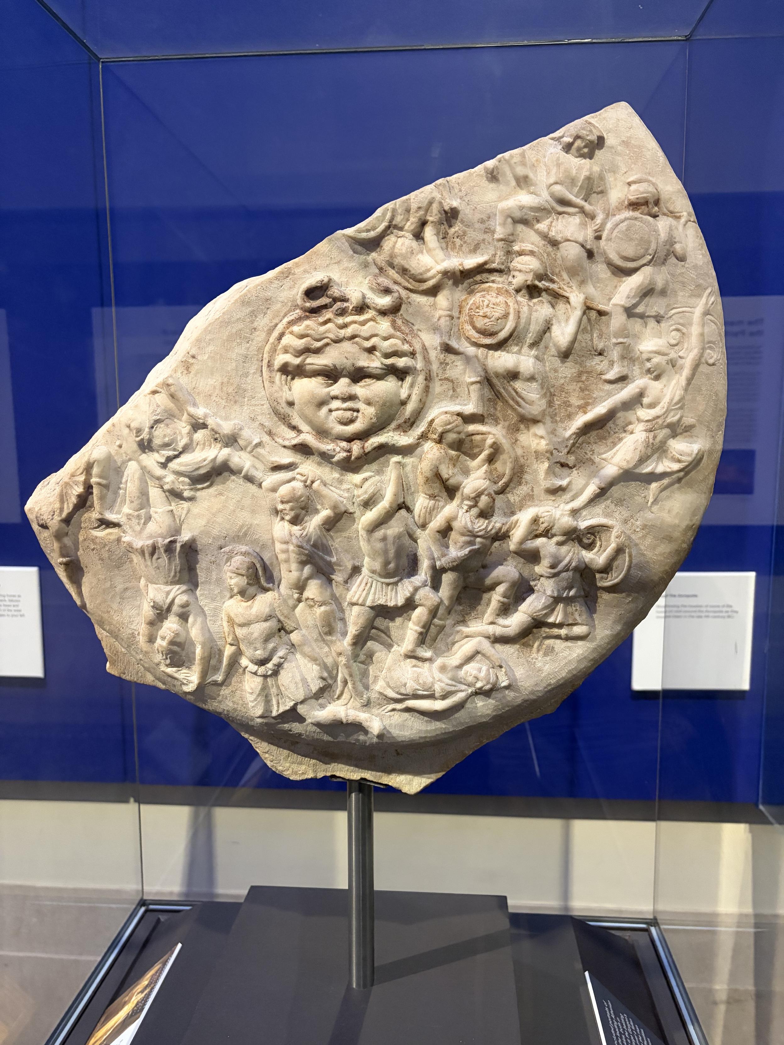 Bas relief avec des guerriers et un gros visage dans un m&eacute;daillon, qui peut faire penser au soleil dans les teletubbies - c&rsquo;&eacute;tait un visage de b&eacute;b&eacute; dans le ciel