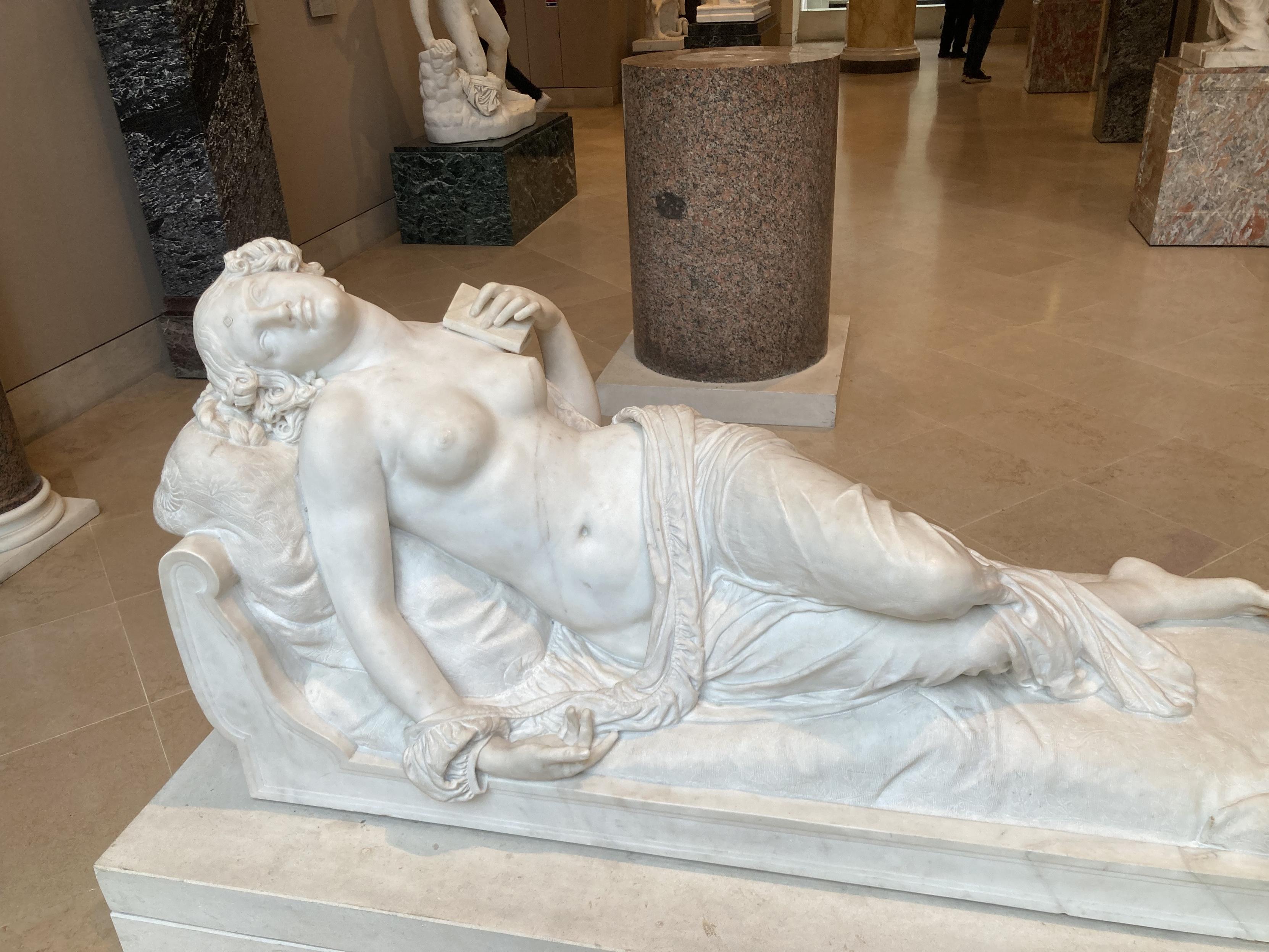 Statue d’une femme allongée dans une pose alanguie, le bas du corps caché par un drap. Elle tient un petit livre dans une main qu’elle a posée sur sa poitrine et à la tête renversée en arrière