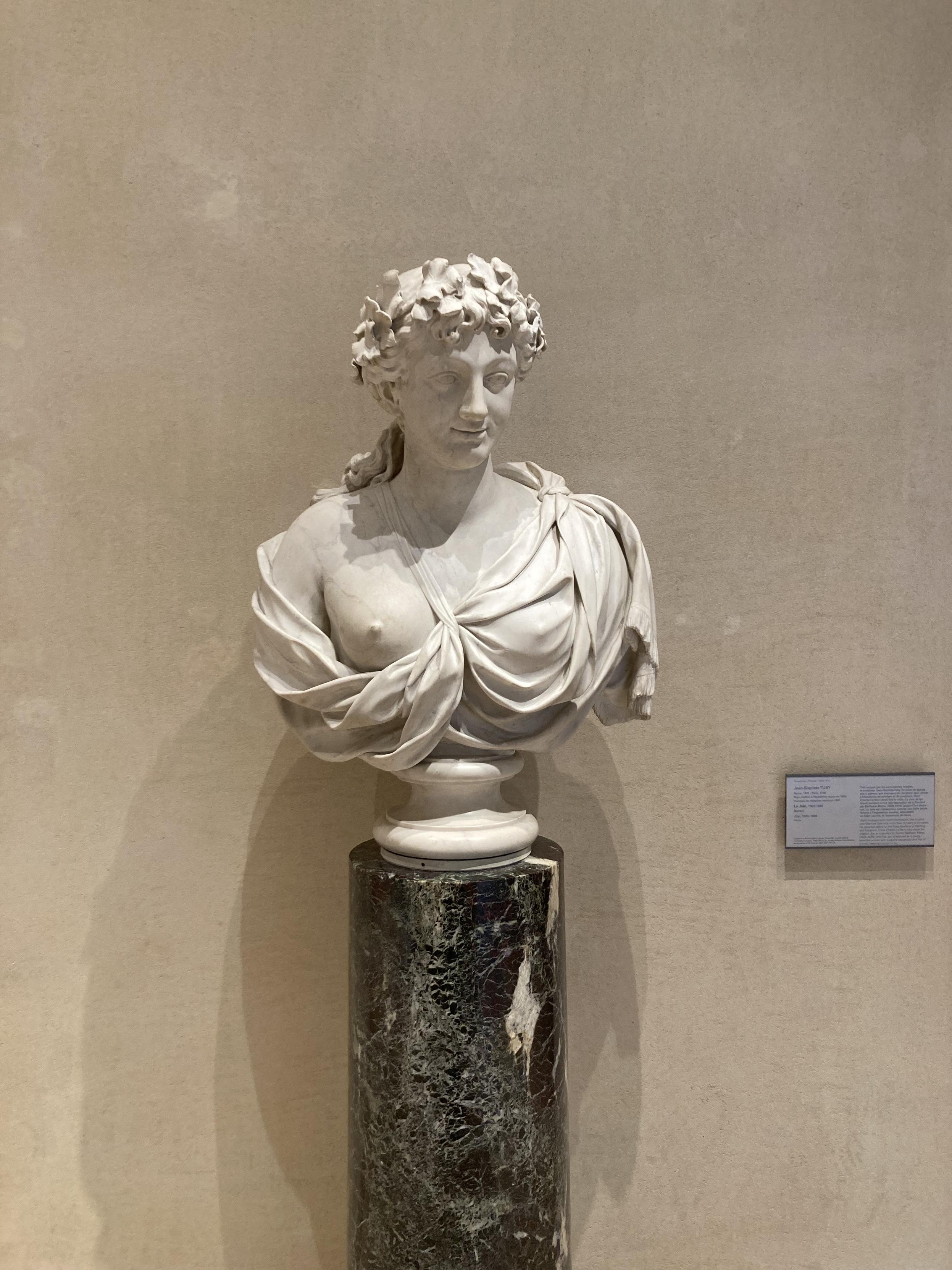 Statue d’un buste de femme, habillé d’un tissu drapé retenu par une bretelle, mais qui laisse voir un de ses seins.