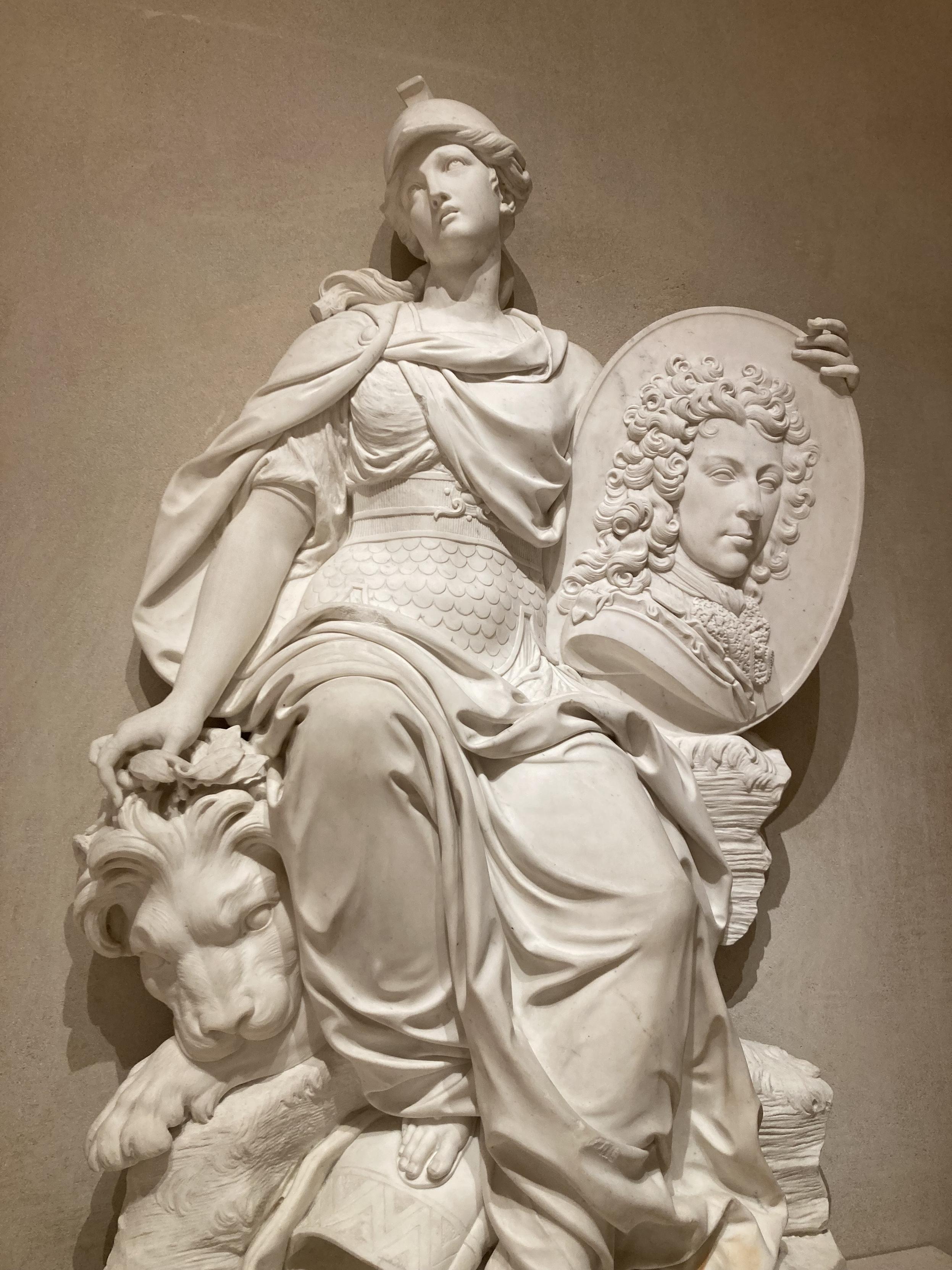 Sculpture d'une femme assiste, tenant un portrait gravé d'un homme à perruque. Elle a l'air turbo saoulée.