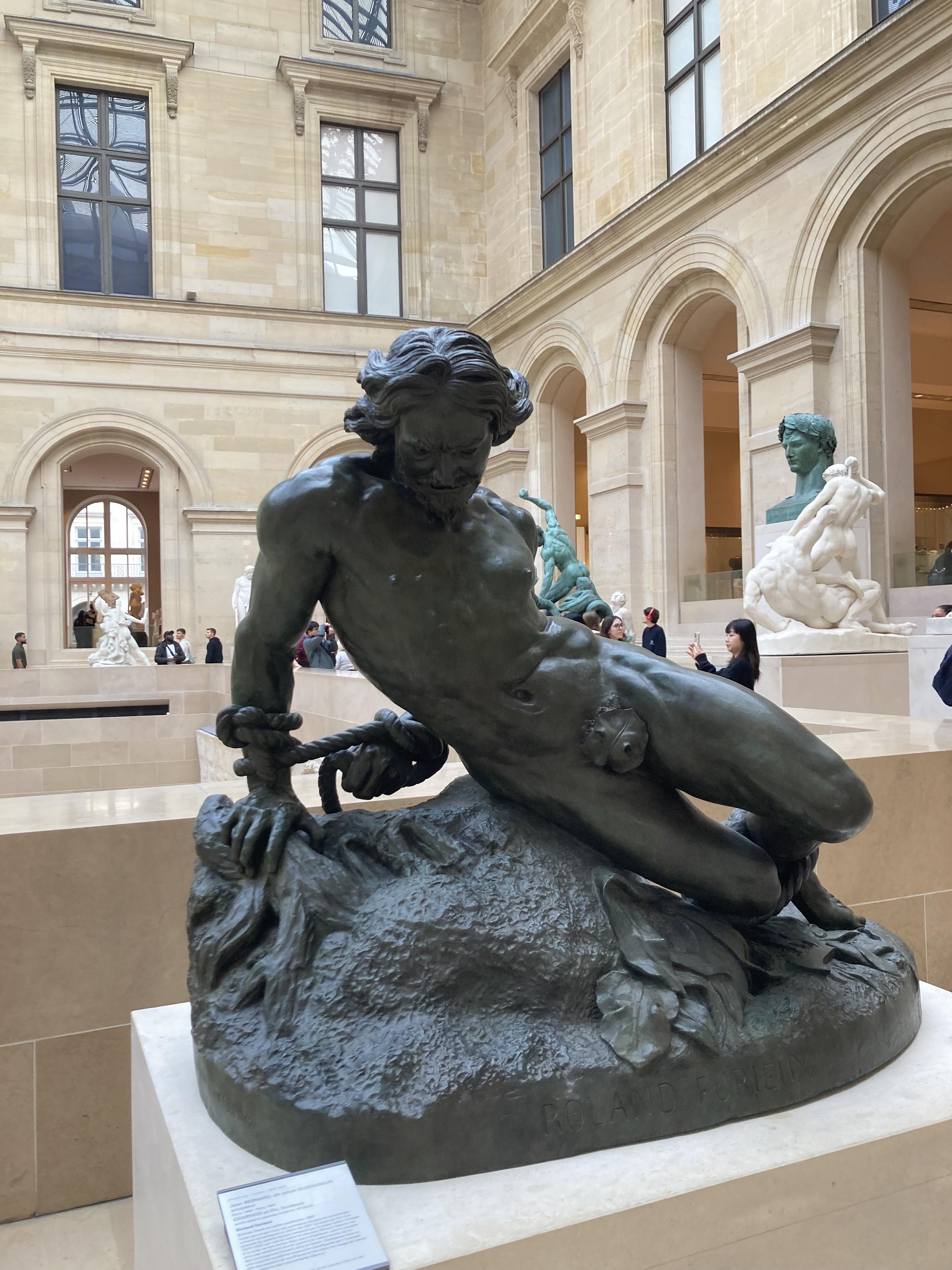 Sculpture d’un homme assis par terre, appuyé sur une main. Ses deux mains sont reliées dans son dos par une corde avec un gros noeud