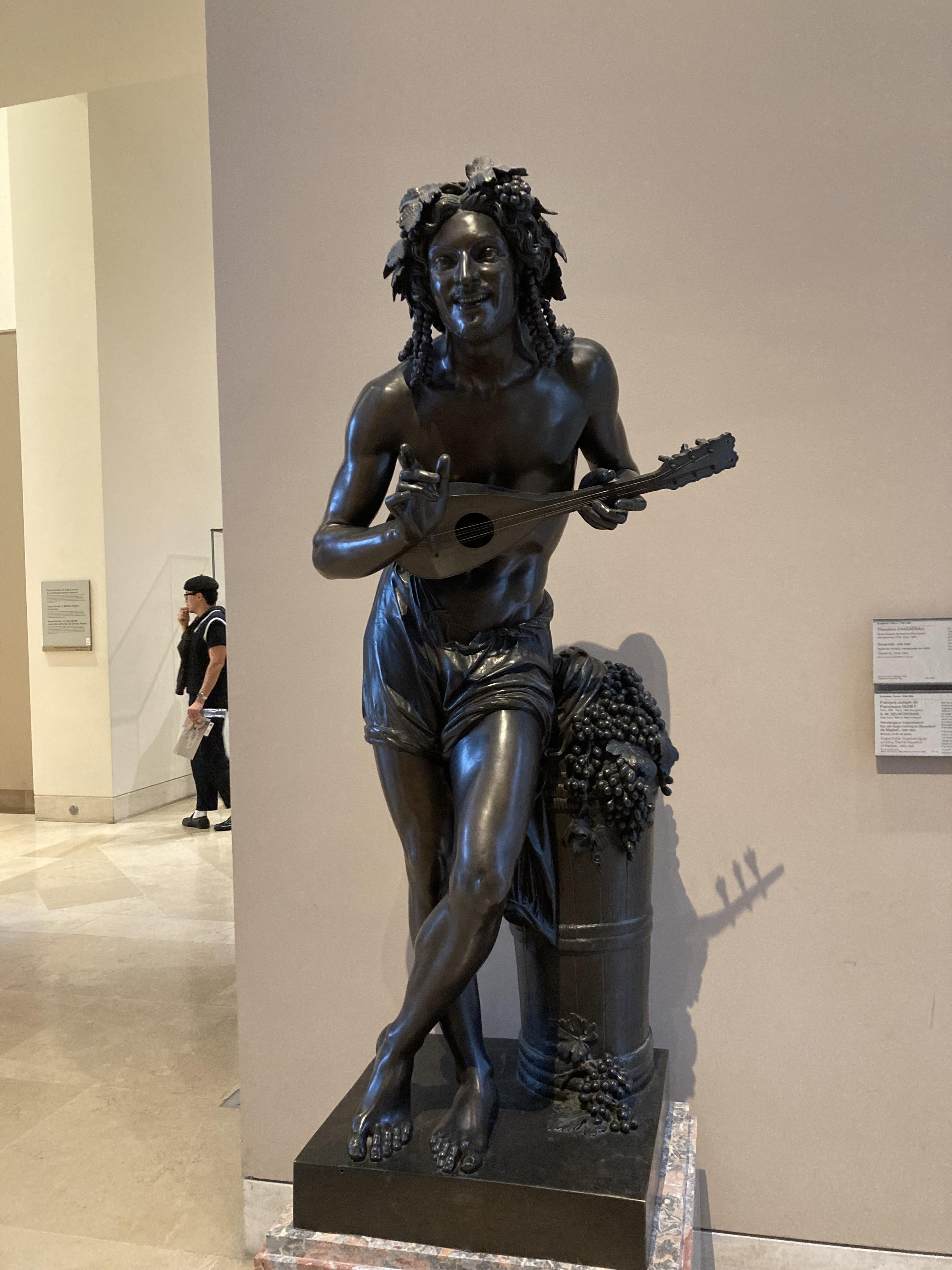 Statue d’un homme debout, en train de jouer de la guitare. Il a les cheveux longs et une couronne de raisin sur la tête, il est torse nu. Il  a une posture très détendue et est légèrement penché en avant, comme s’il était en train de s’adresser à quelqu’un en face de lui
