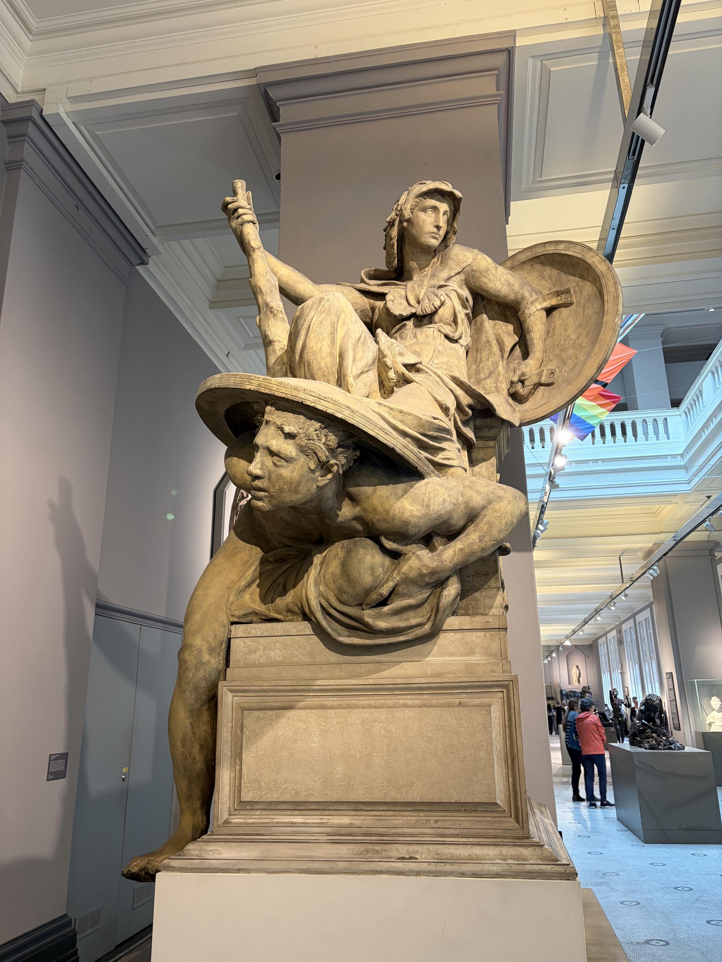Sculpture d&rsquo;une femme assise sur un bouclier, elle m&ecirc;me avec une arme et un bouclier au bras. Sous le bouclier, &eacute;cras&eacute;, un homme.